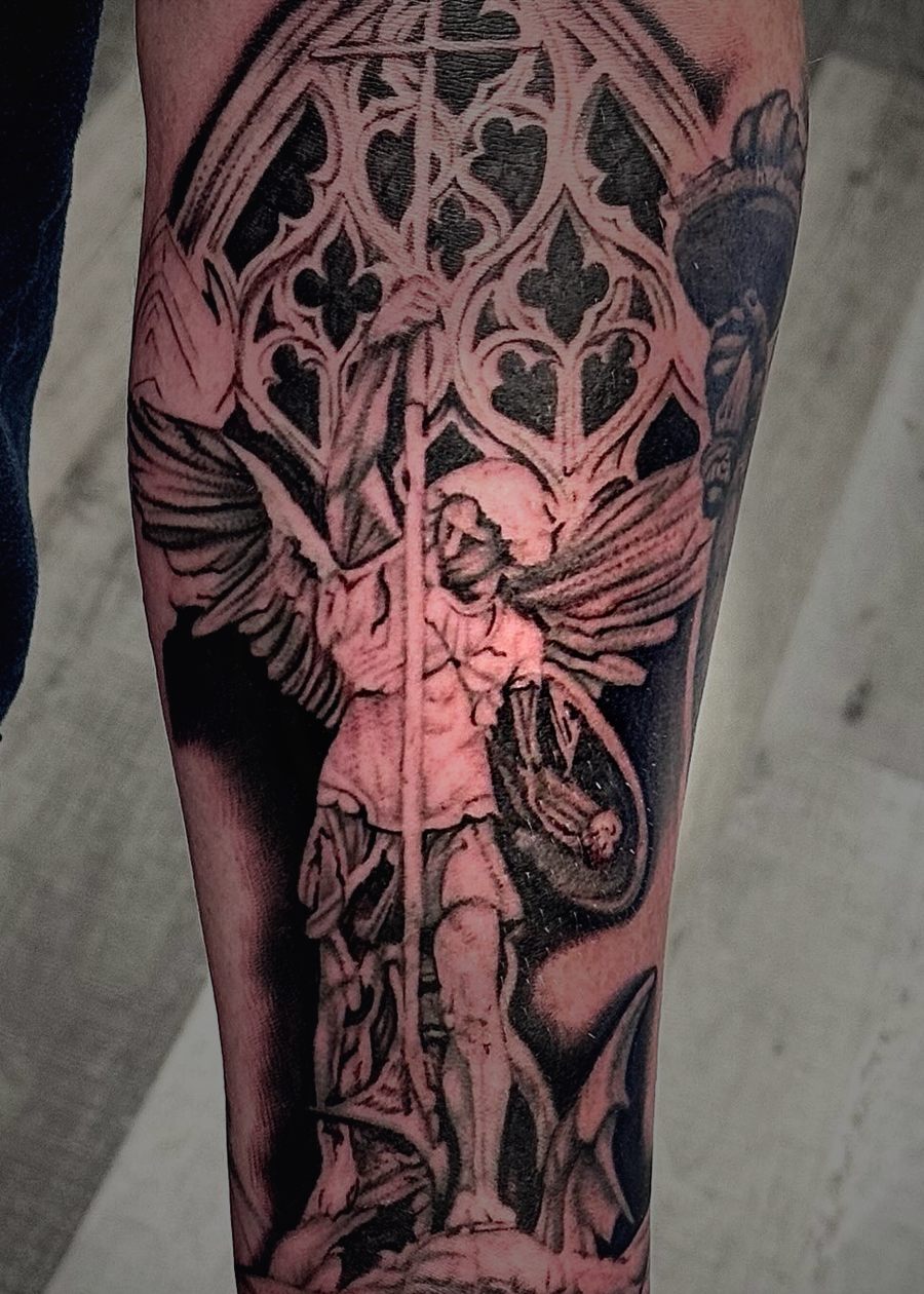 Black and grey archangel standing over a demon under a gothic window, on a forearm.
