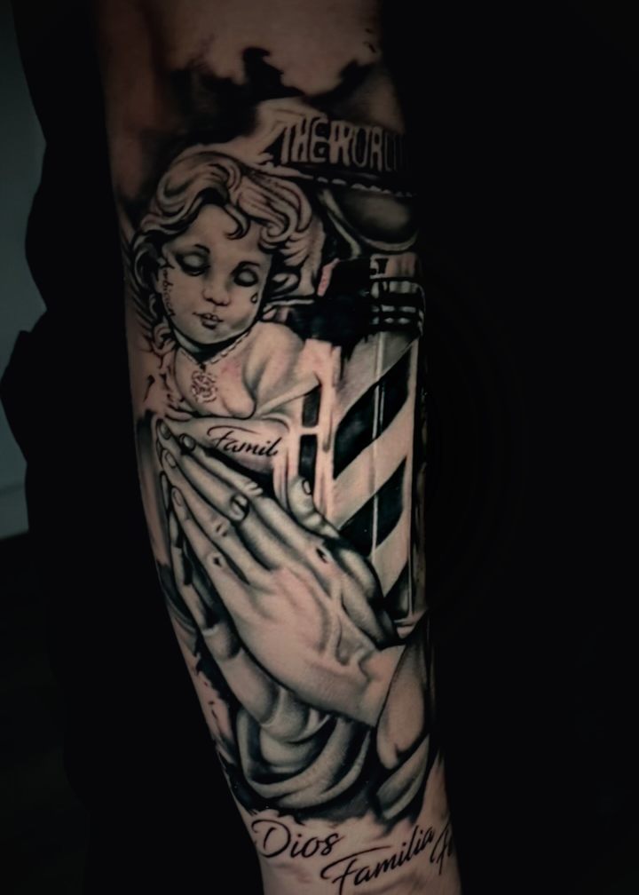Black and grey chicano style cherub with praying hands and script lettering on a forearm.