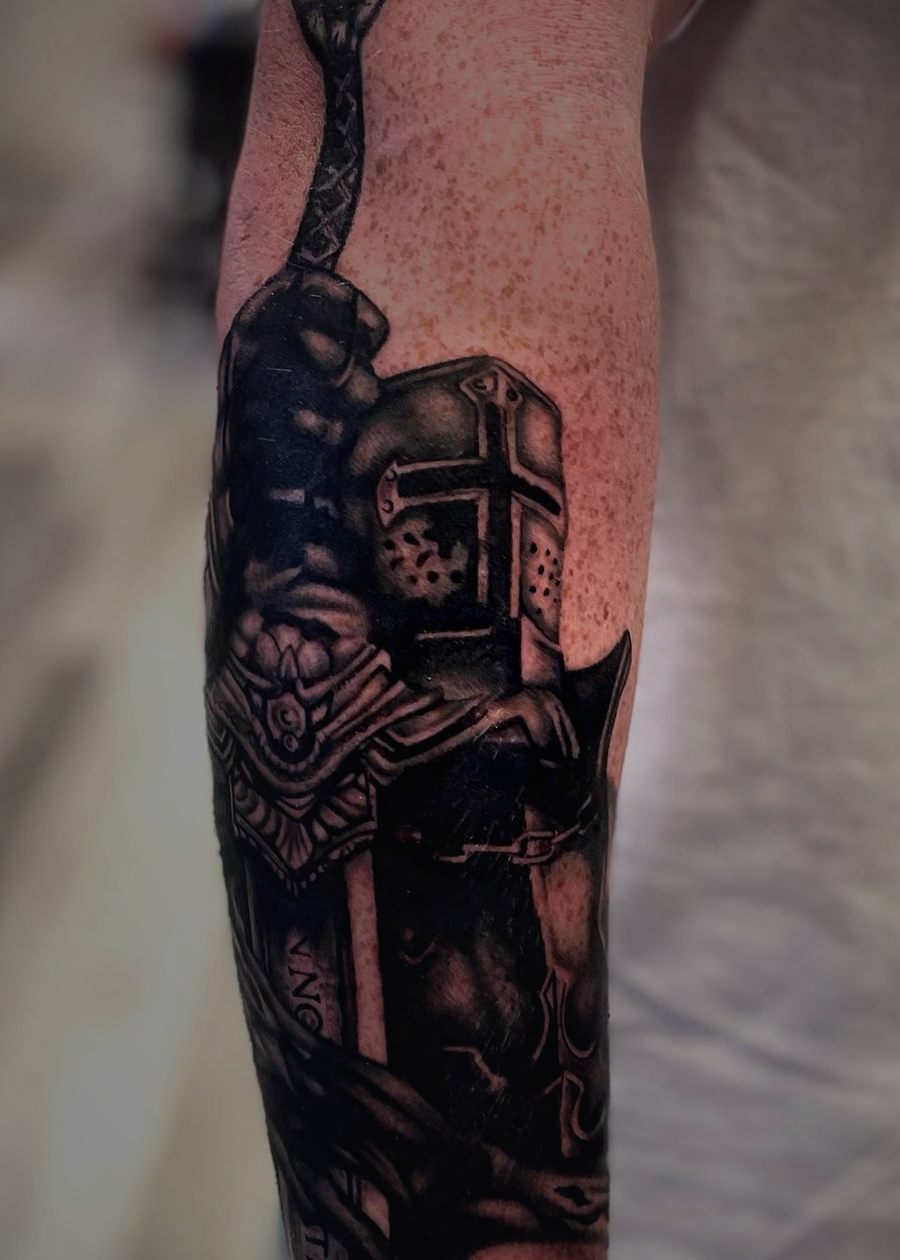 Black and grey crusader in a helmet holding a sword, tattooed on a forearm.