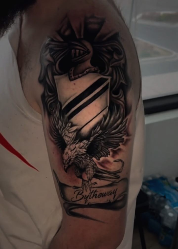 Black and grey crest with a knight's helm, shield and an eagle over a lettered banner, on a shoulder.