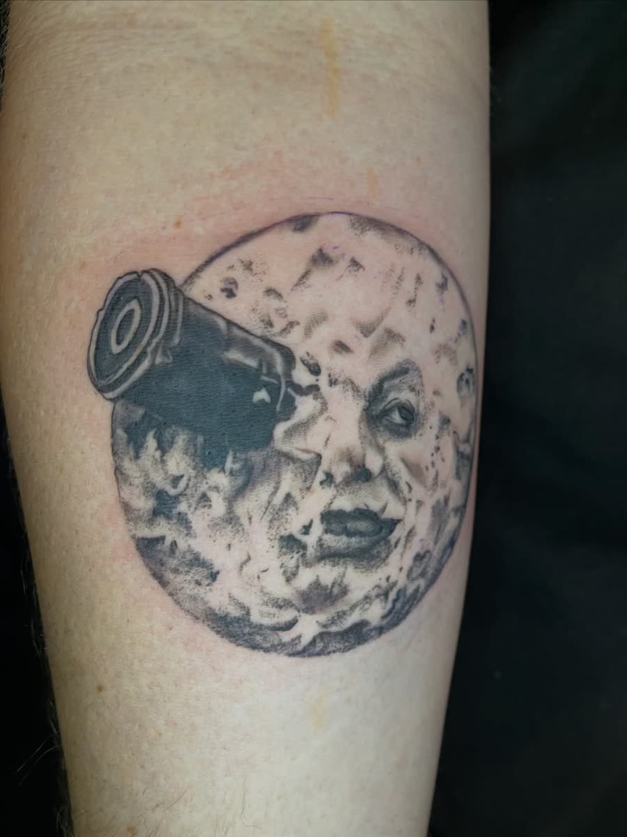 Black and grey moon face with a rocket in its eye, tattooed on a calf.