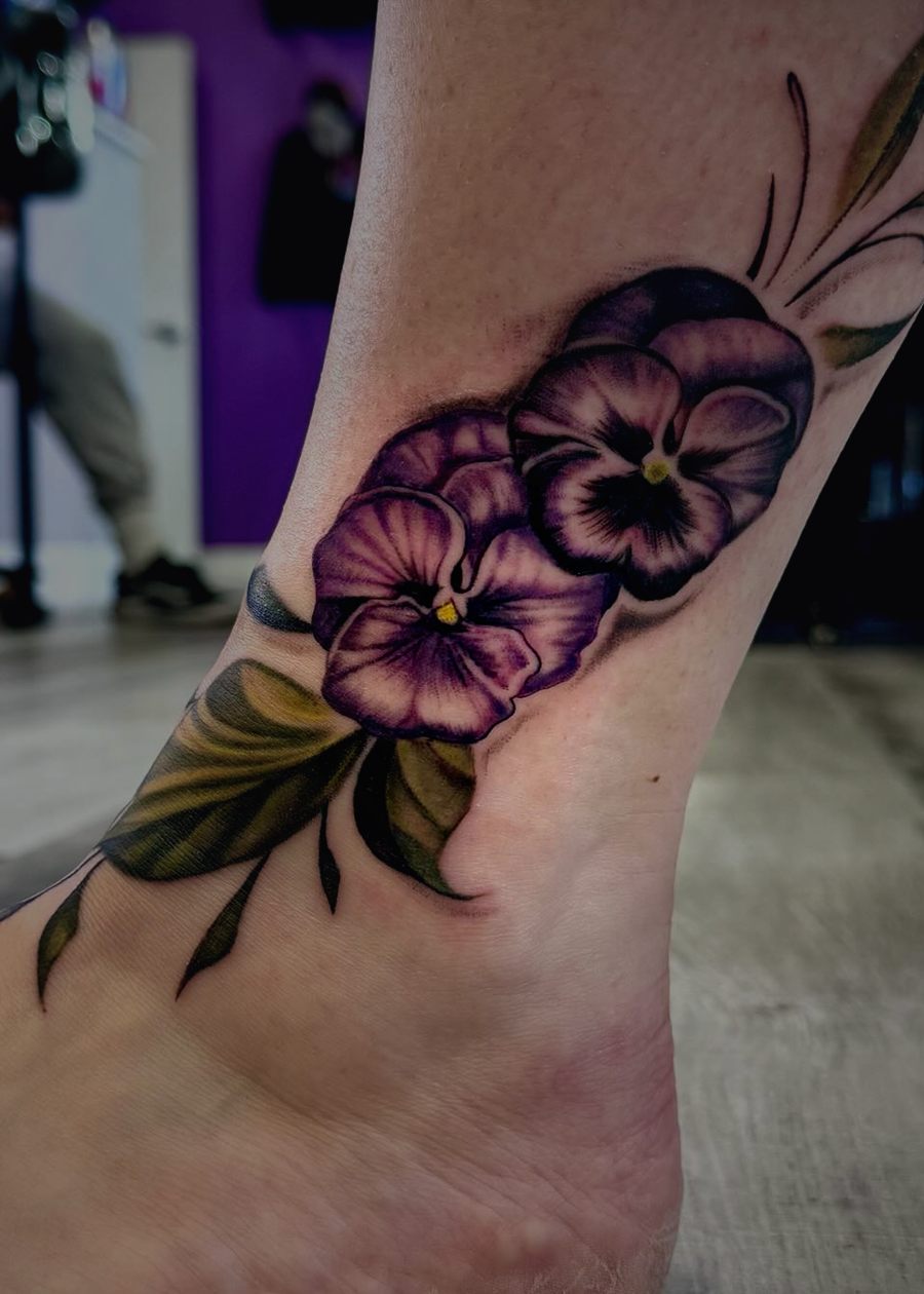 Purple pansy flowers with green leaves tattooed around an ankle.