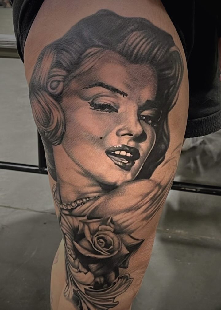 Black and grey pin-up portrait with a rose beneath it, tattooed on a thigh.